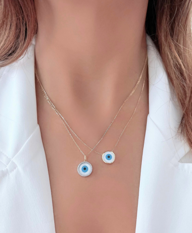 A person wearing a white shirt showcasing two necklaces, one with a round mother of pearl charm and a smaller evil eye charm, both with gold trim, against their neck.